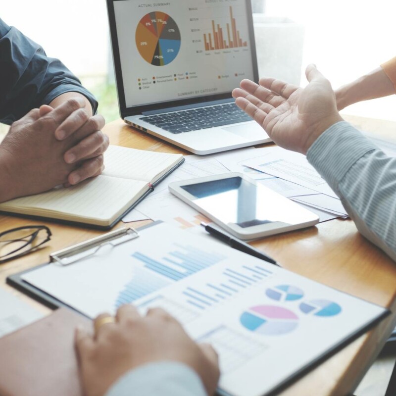 Two people sit at a desk surrounded by multiple spreadsheets on a laptop and on the desk. These spreadsheets are made more efficient with the help of AI.