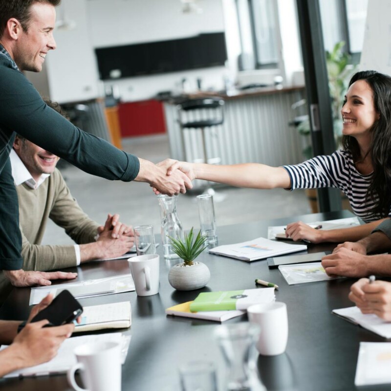 A group of employees sitting at a desk onboarding a new employee. The new hire extends his hand to shake the hand of his new team member.