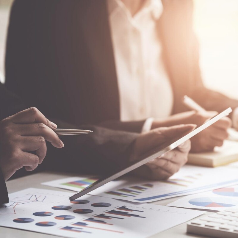Two people sitting at a desk holding tablets and papers of graphs. An executive administrative assistant is supporting their executive in performing their job duties optimally.