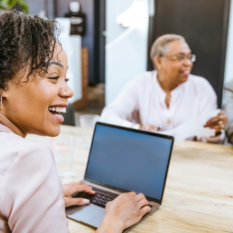 An administrative assistant sitting in on a meeting smiling with her coworkers. She's sitting at her laptop, demonstrating how to take meeting minutes effectively.