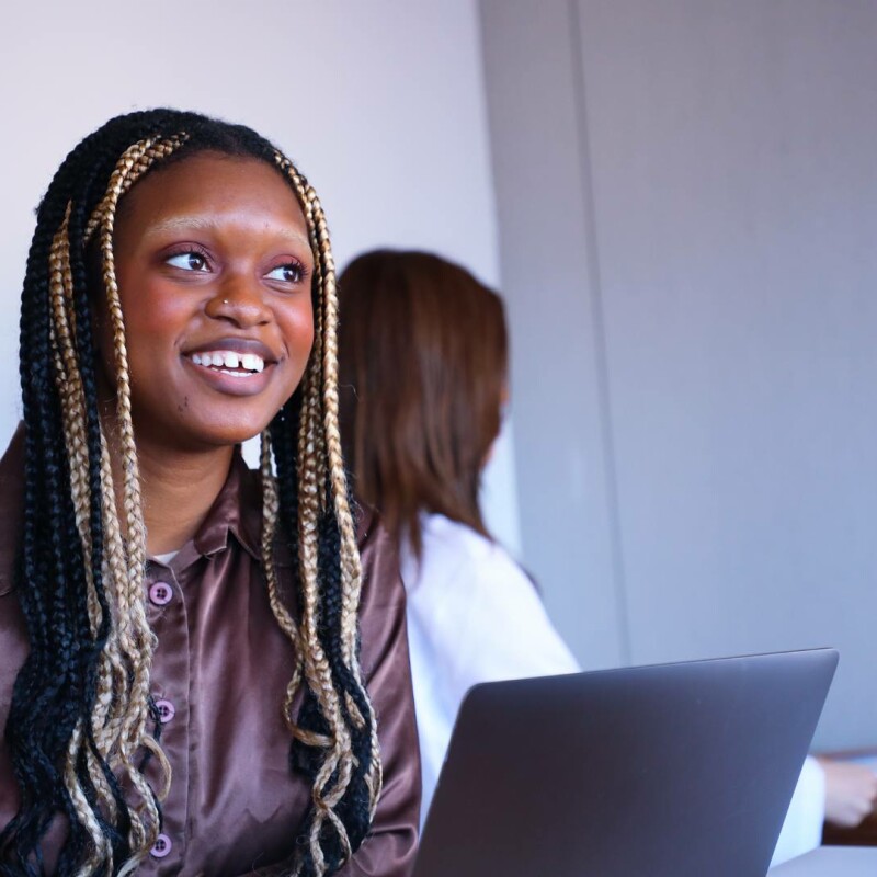 An administrative assistant sits at a desk with an open laptop, speaking with a coworker.