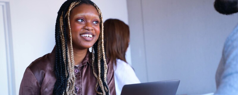 An administrative assistant sits at a desk with an open laptop, speaking with a coworker.