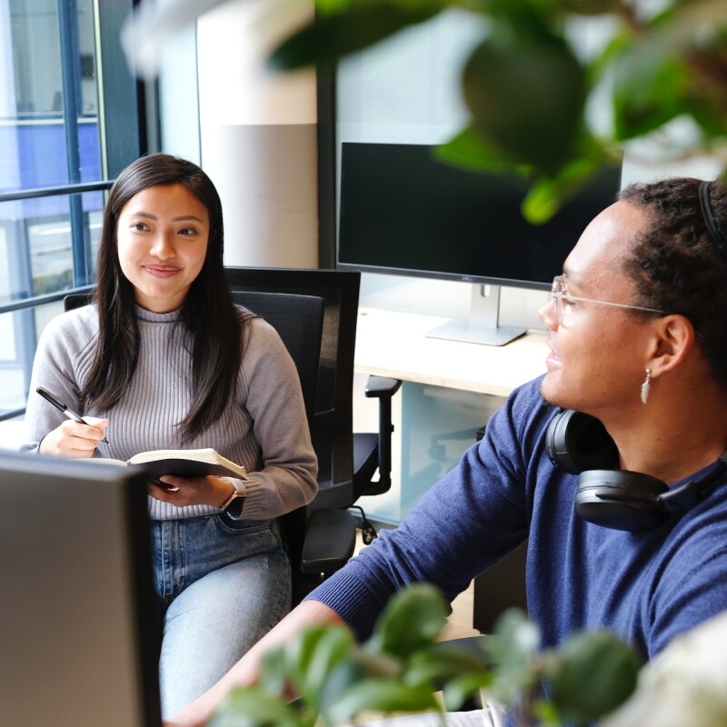 An administrative or executive assistant writes in notebook, sitting next to coworker who is working at a computer
