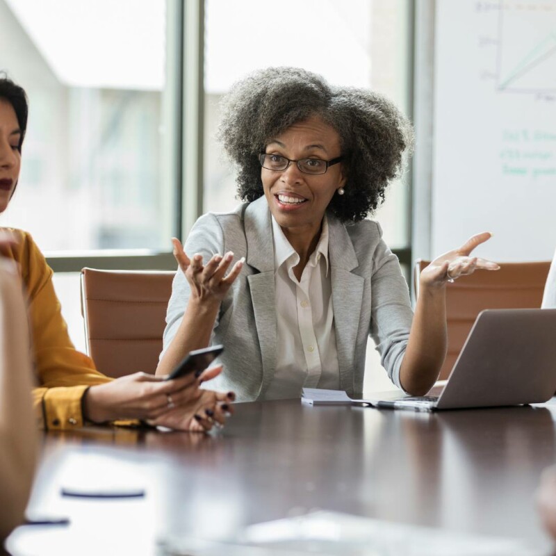 Three women sit around a conference room table with a laptop, cellphone, and binder. They are part of the board of directors.
