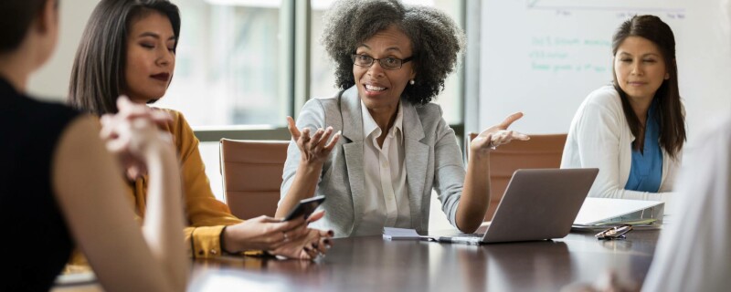 Three women sit around a conference room table with a laptop, cellphone, and binder. They are part of the board of directors.