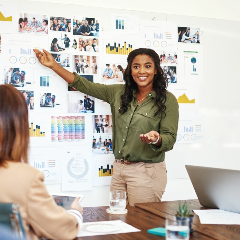 A woman stands in front of her colleagues, pointing at images and points on a whiteboard as they plan. Image demonstrates how to build trust at work.