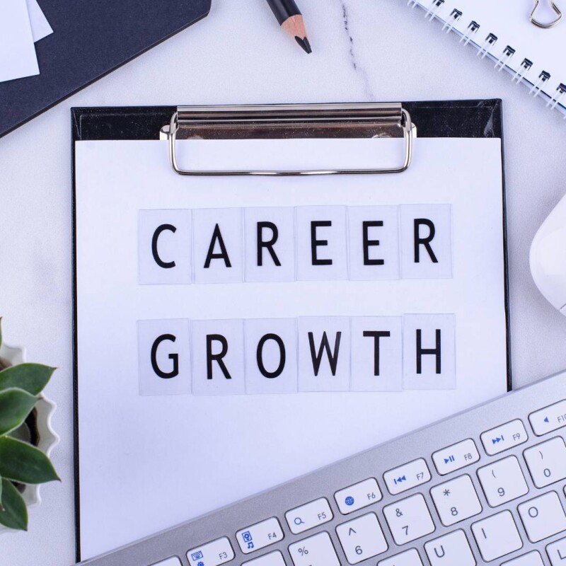 A clipboard with the words "career growth" on it is displayed on a desk surrounded by a keyboard, mouse, and green plant.