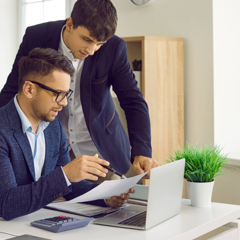 One person sits at a desk and another person stands behind them. Both of them are wearing suits. They're viewing a laptop for content management.