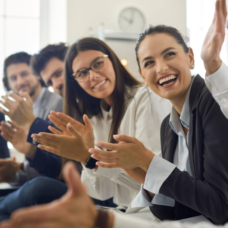 A group of employees sits together while smiling and clapping at a fellow employee, demonstrating employee appreciation.