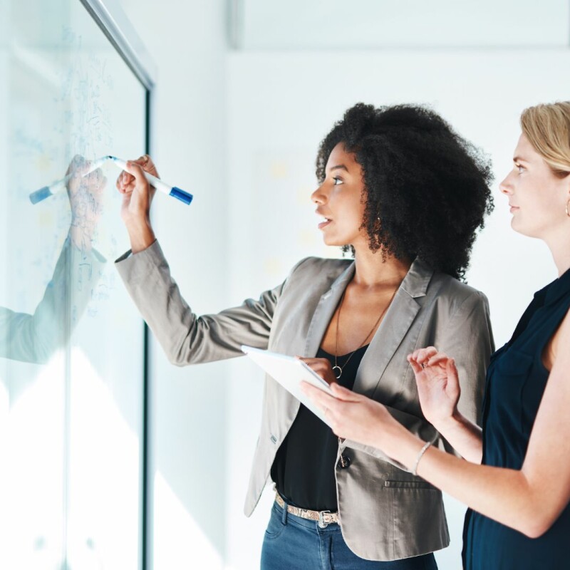 Two women stand in front of a see-through whiteboard, and one of them is writing things down, explaining to the other. Image demonstrates an employer supporting employee development.
