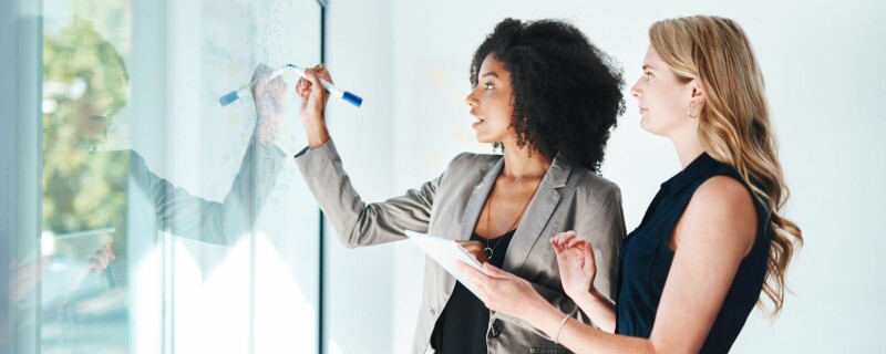 Two women stand in front of a see-through whiteboard, and one of them is writing things down, explaining to the other. Image demonstrates an employer supporting employee development.