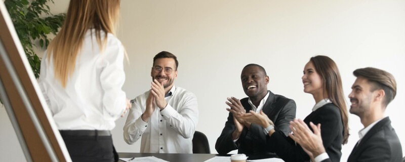 A group of four employees sit and clap as they look at their boss, who stands in front of them. They are demonstrating employee appreciation.