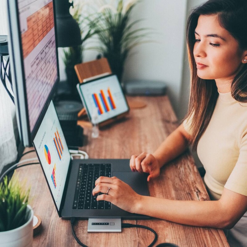 An executive assistant sits at her desk and looks as graphs on a laptop and desktop computer, demonstrating KPI management.