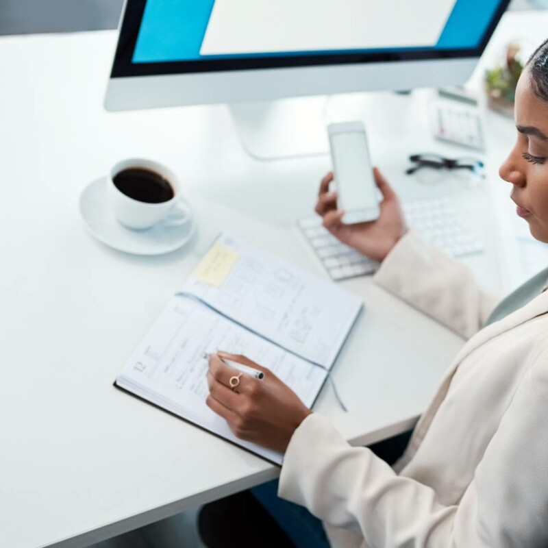 An executive assistant sits at her desk holding her phone while writing in her planner. A cup of coffee sits in front of the planner.