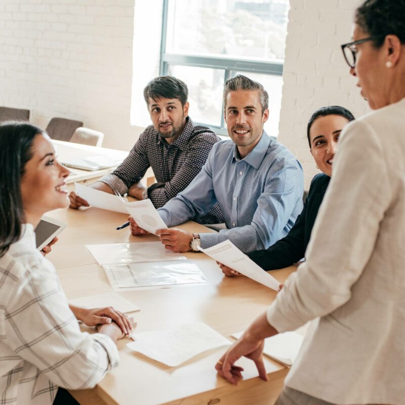 A group of six employees sit around a table and listen to their boss speak. Their boss is displaying executive presence.