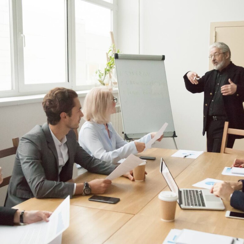 An administrative assistant stands at the front of a conference table, demonstrating leadership skills to fellow employees.