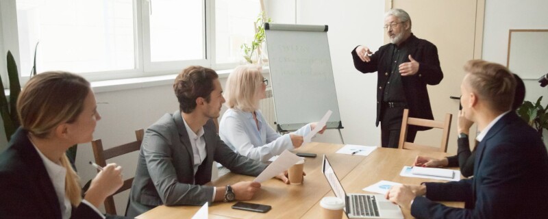 An administrative assistant stands at the front of a conference table, demonstrating leadership skills to fellow employees.