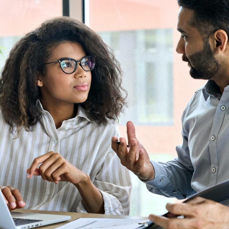 Two people, a mentor and mentee, sit at a desk and chat with one another. There is a green leafy plant in the background and a silver computer on the desk.