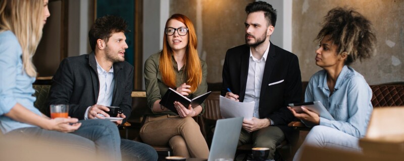 A group of coworkers sit together in the workplace, and a few of them look slightly offended by what another is saying.