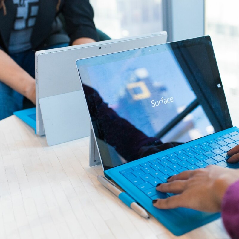 Two coworkers sit across from each other on a table, working on Microsoft Tablets. Image demonstrates how Microsoft OneDrive can be used collaboratively.
