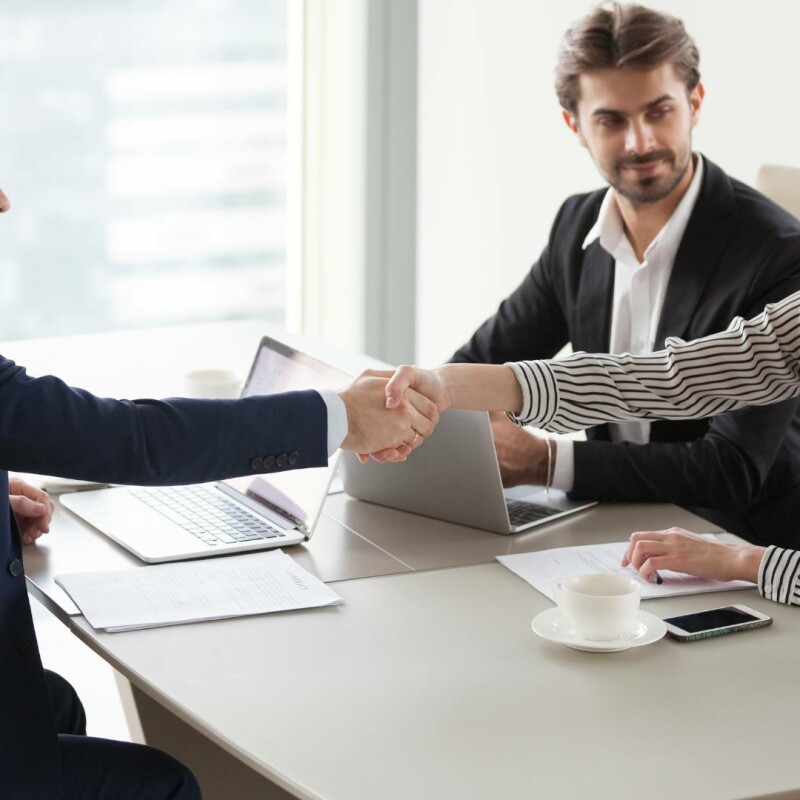 Two men and one woman sit around an office desk. The desk has two laptops, a cell phone, paperwork, and a white coffee cup on it. One man and the woman shake hands to seal the deal on a corporate venue negotiation.
