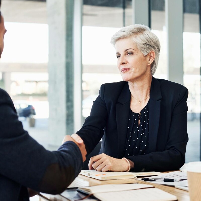 Two people sit at a table in a conference room and shake hands, demonstrating a successful negotiation.