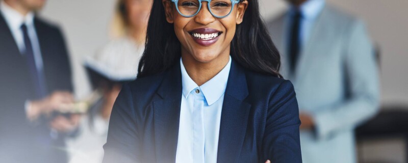 A woman stands in front of the camera wearing glasses and smiles. There are three other people standing behind her. The photo demonstrates office professionals and personal branding.