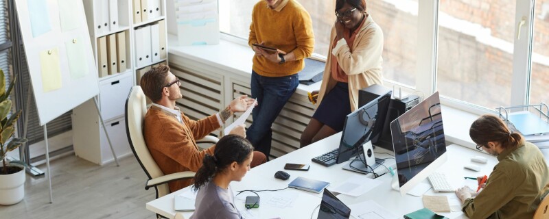 A group of coworkers are sitting and standing around a table, working together. Image demonstrates the importance of measuring productivity.