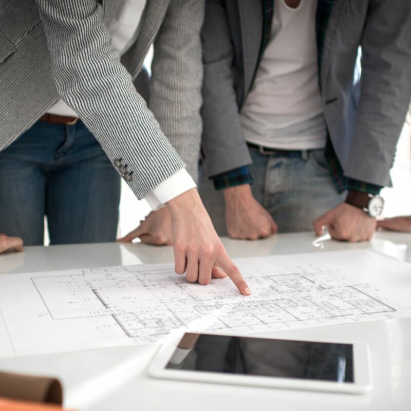 A group of coworkers gather around their project manager at the table, looking over blueprints.