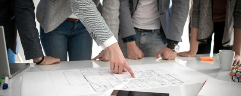 A group of coworkers gather around their project manager at the table, looking over blueprints.