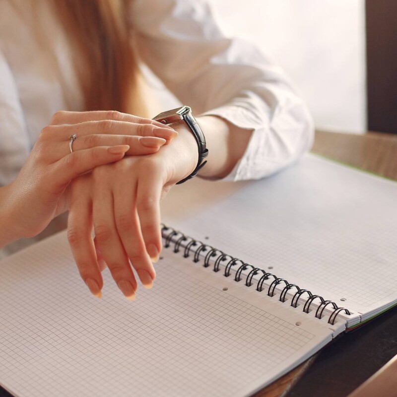 An administrative employee sits at a desk and checks their watch over their notebook, demonstrating time management.