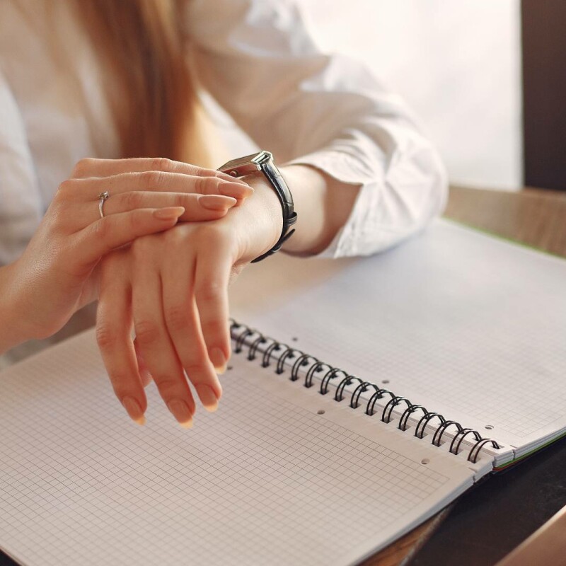 A person looks at their watch at their desk with an open notebook beneath, demonstrating time management skills.