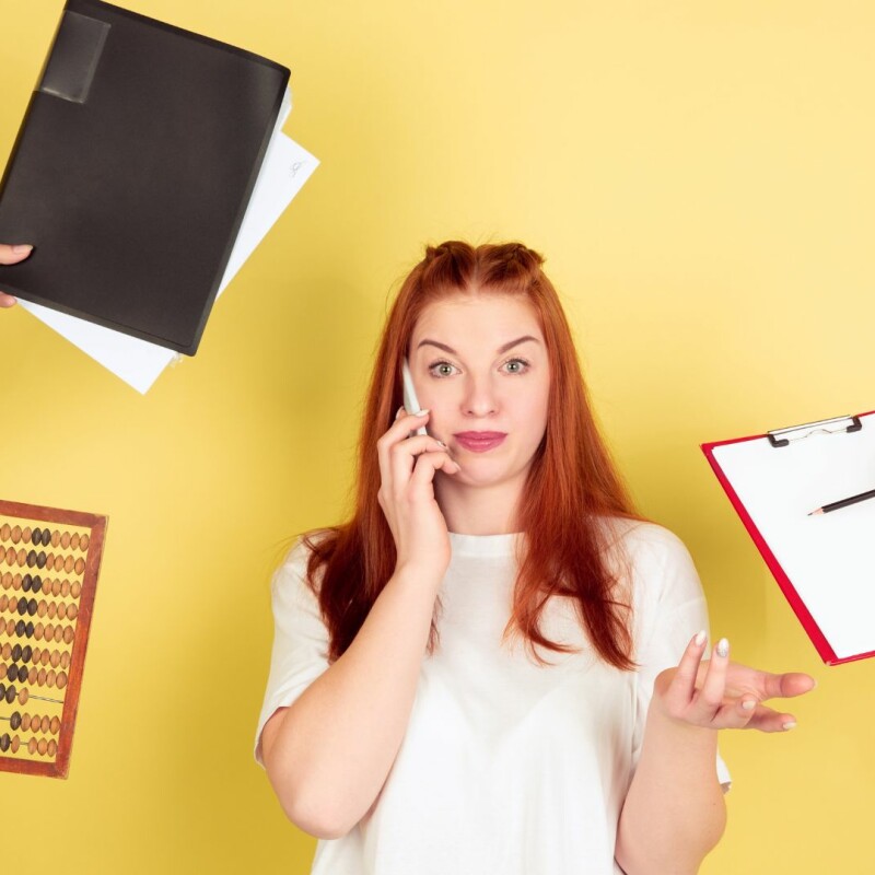 A woman stands in the middle of the photo with a phone pressed against her ear. Several hands are coming in from off-screen, trying to give her a notebook, a clipboard, and an abacus. Image demonstrates the need for time management skills.