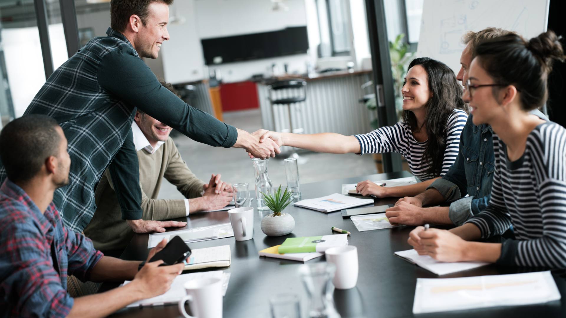 A group of employees sitting at a desk onboarding a new employee. The new hire extends his hand to shake the hand of his new team member.