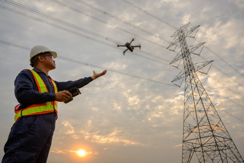 Asian male engineer electrician at power station flying drone to observe power generation planning work at high voltage pylons