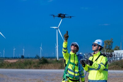 Two engineers in safety gear operate a drone to survey a wind farm, showcasing modern technology in renewable energy maintenance and inspection.