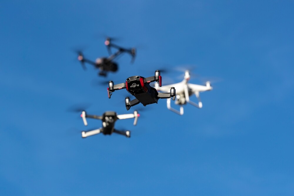 A diverse fleet of drones of all sizes and weights soars through a clear blue sky, their sleek frames glinting in sunlight as they maneuver gracefully in synchronized flight