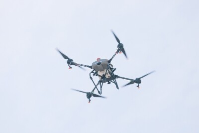 Close-up view of a modern agricultural drone in flight against a bright sky. The drone is equipped with spraying nozzles and sensors used for precision farming, crop monitoring, and agricultural.