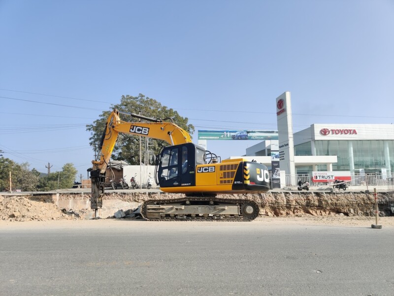 A robust yellow JCB excavator stands ready at a roadside construction site, set against a backdrop of a clear blue sky.