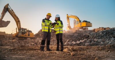 Caucasian Male Civil Engineer Talking To Hispanic Female Inspector And Using Tablet On Construction Site of New Apartment Complex.