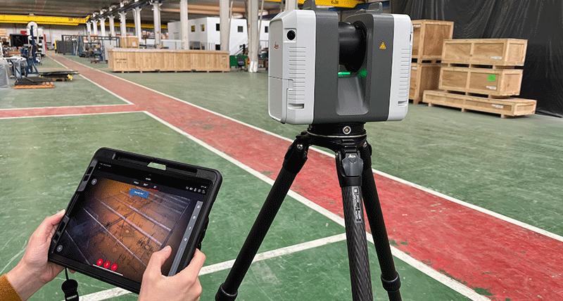 Laser scanner on a factory floor with person holding a tablet displaying scan data.