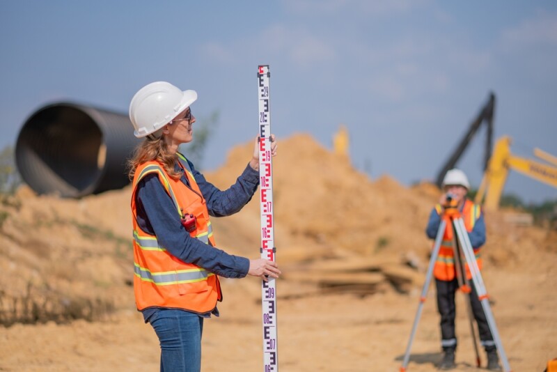 Construction surveyor or engineer working construction site leveling rod and theodolite mounted tripod measuring angles and distances with heavy machinery excavator constructed hi-way road background