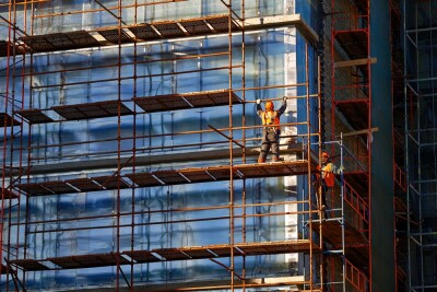 Worker installing scaffolding on building, showcasing the teamwork and safety equipment required for scaffold setup on construction site, focusing on safety harnesses and work platforms.