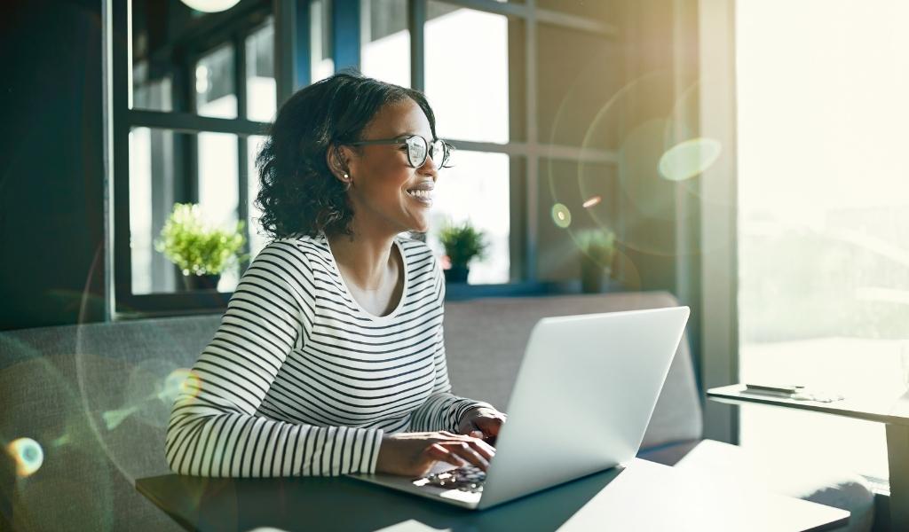 Woman smiling at laptop while working while the sun shines in
