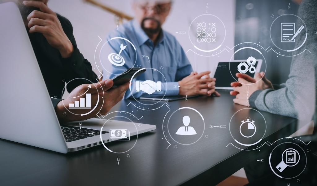Colleagues in a meeting with icon imagery overlaid containing: target, graph, clock, cash, gears