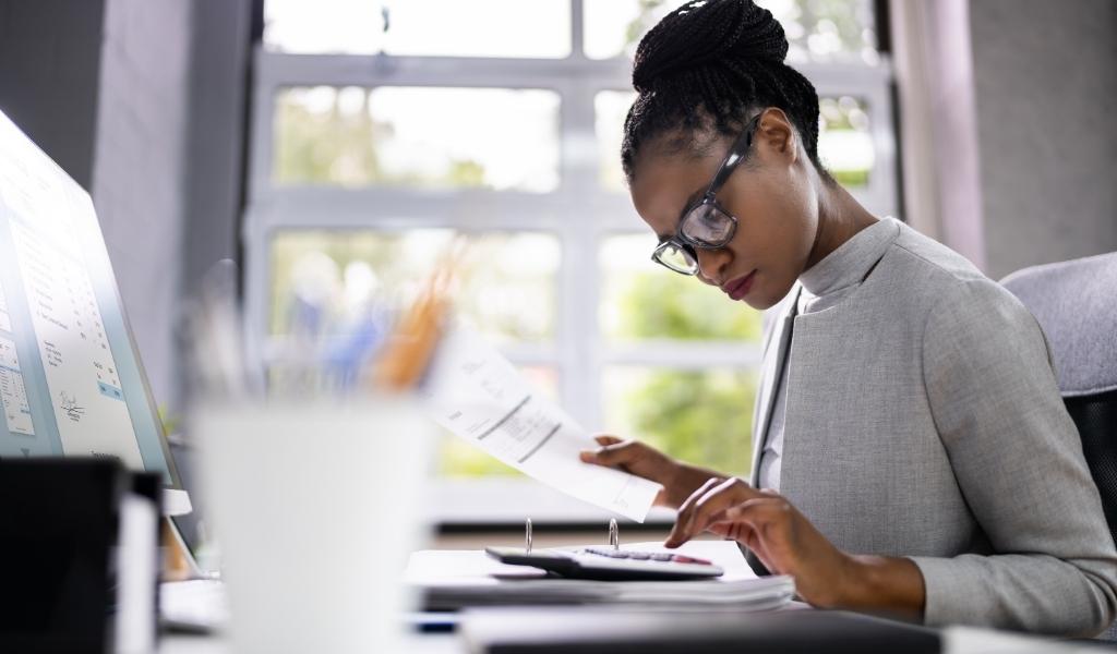 Woman working at computer while using calculator and holding paper