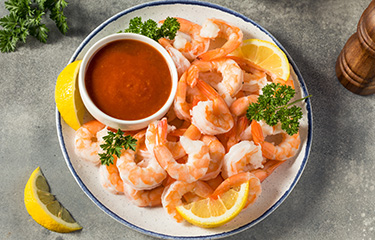 A plate of cooked shrimp with a dipping sauce and lemon slices.