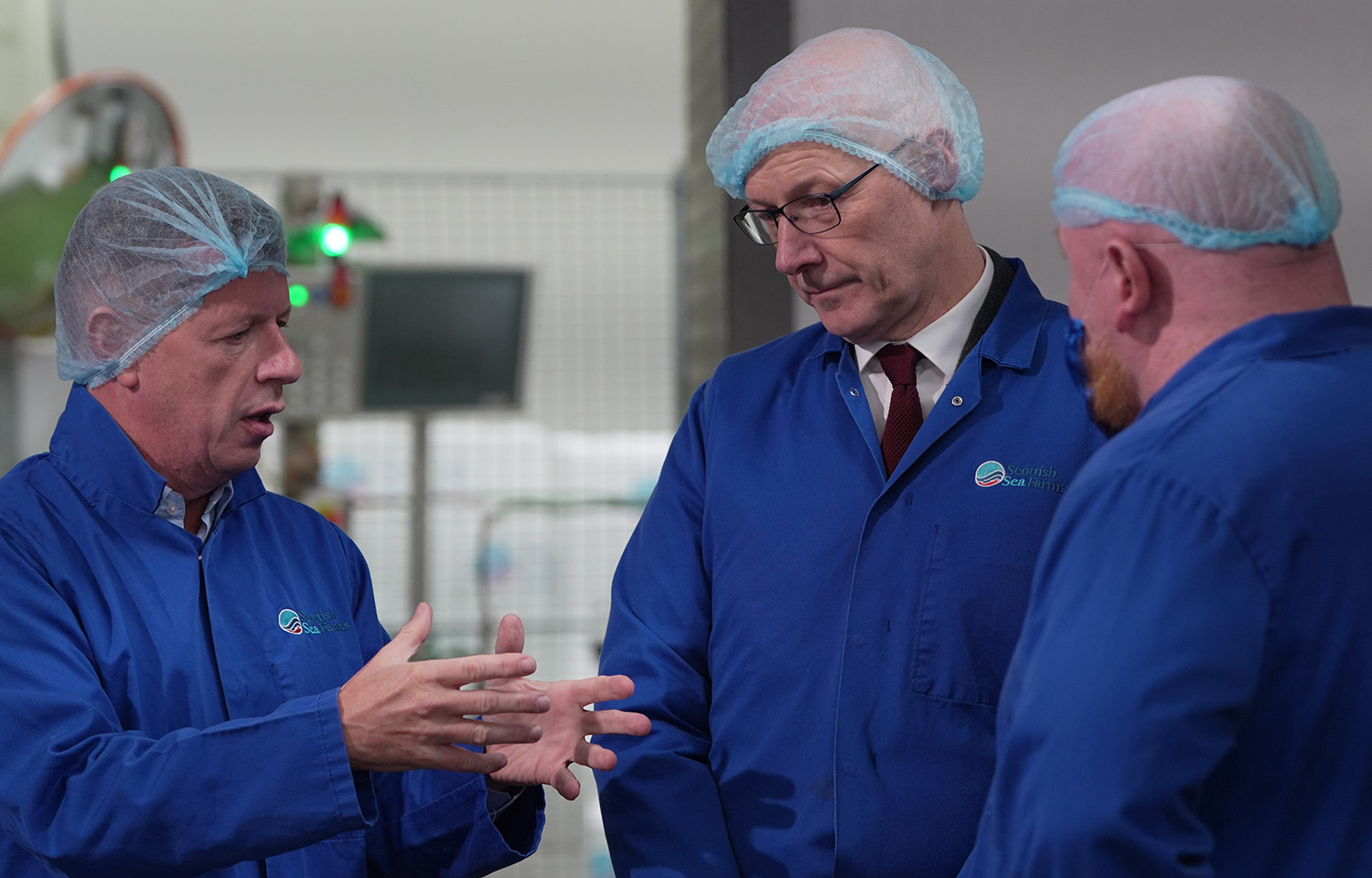 Scotland First Minister John Swinney in a hairnet and jacket talking with Scottish Sea Farms employees in a processing facility
