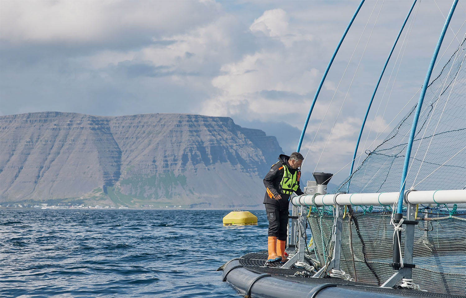 An Icelandic Salmon employee standing on the edge of a salmon net pen with mountains in the background.