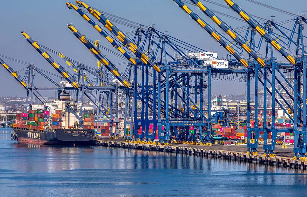Blue and yellow cranes suspended over the port of Los Angeles, where a blue container ship waits, stacked high with multicolored containers.